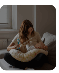woman sitting cross-legged with a pillow on her legs and a baby on the pillow, shaking a rattle and smiling at her baby