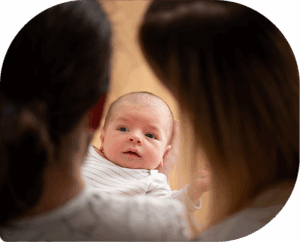 Newborn baby, seen from behind and over the shoulder of the woman who's holding him and another woman who's also watching the baby