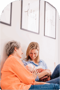 A grandmother and a mother and her baby, sitting on the floor, leaning against a wall comfortably, smiling while looking at the baby
